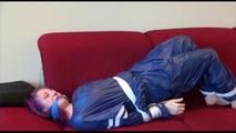 Mara tied and gagged on a sofa wearing a shiny blue PVC sauna suit (Video)