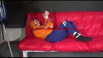 Lucy wearing a blue rain pants and an orange rain jacket being tied and gagged with belts on a sofa (Video)