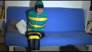 Mara tied, gagged and hooded with tape on a blue sofa wearing an blue/green rain combination (Video)
