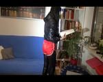 Mara wearing a red shiny nylon shorts over a black rainpants and a rain jacket while searching a book and lolling on the sofa (Video)