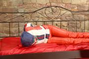 Mara tied, gagged and hooded on bed wearing a shiny red/blue/white striped old school down jacket and a skibib in red (Pics)