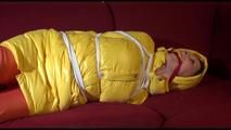 Lucy tied, gagged and hooded on a red sofa wearing an orange venice beach pant and a yellow down jacket (Video)