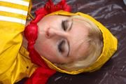 Pia tied and gagged in bed in a yellow rainsuit and a red gag